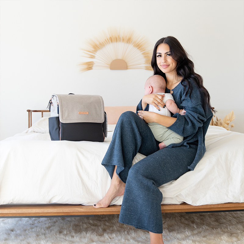 Mother sitting on bed, holding baby, next to a Petunia Pickle Bottom Diaper Bag