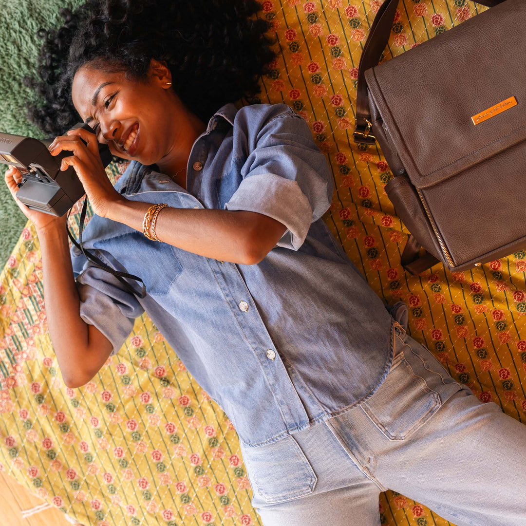 A woman lying on a blanket on the grass taking a photo of whats in front of her. The Boxy Backpack in Saddle is lying face up on the blanket next to her. 