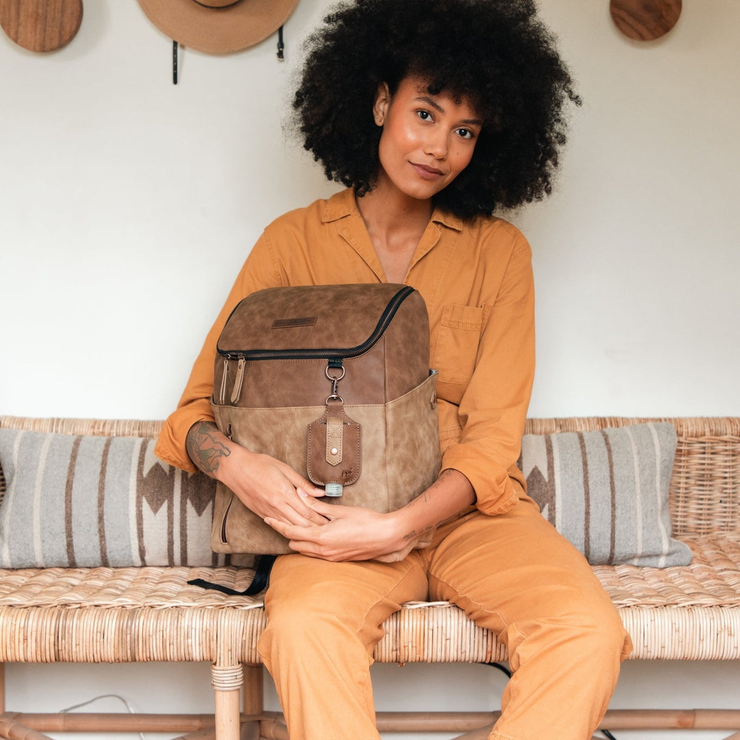 Woman staring at the camera holding a Petunia Pickle Bottom bag in Brioche. She is wearing an orange shirt and orange pants and is sitting on a brown bench against pillows. 