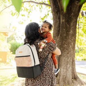 mom wearing inter-mix compatible diaper bag axis backpack holding toddler daughter standing under tree on sidewalk