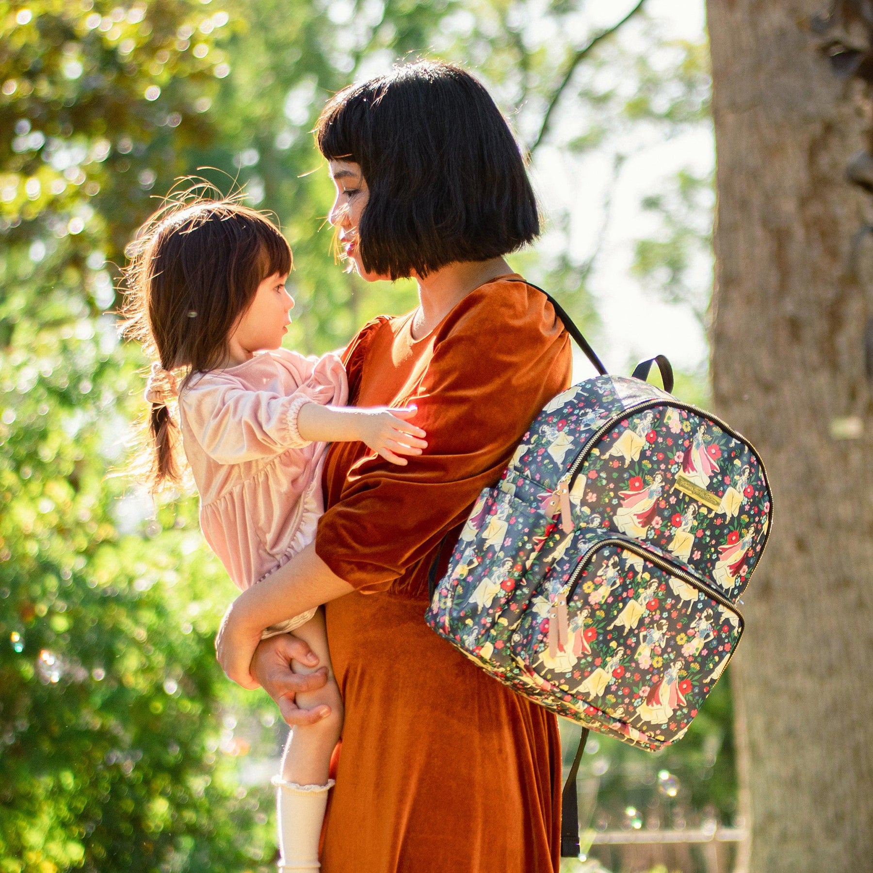 Mom in red dress, looking at daughter in pink dress, holding Disney's Snow White's Enchanted Forest District backpack