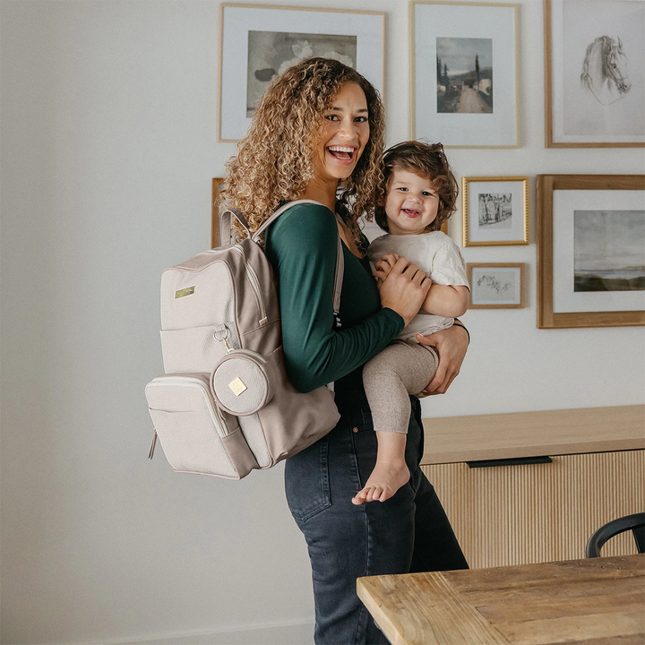 mom carrying baby. petite porter in grey matte cable stitch is attached to d ring of sync backpack in grey matte cable stitch. 