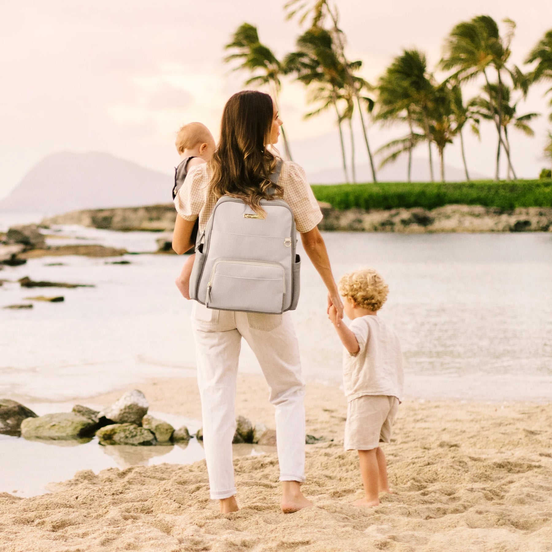 mom wearing the sync backpack in grey matte leatherette while holding baby and hand of toddler