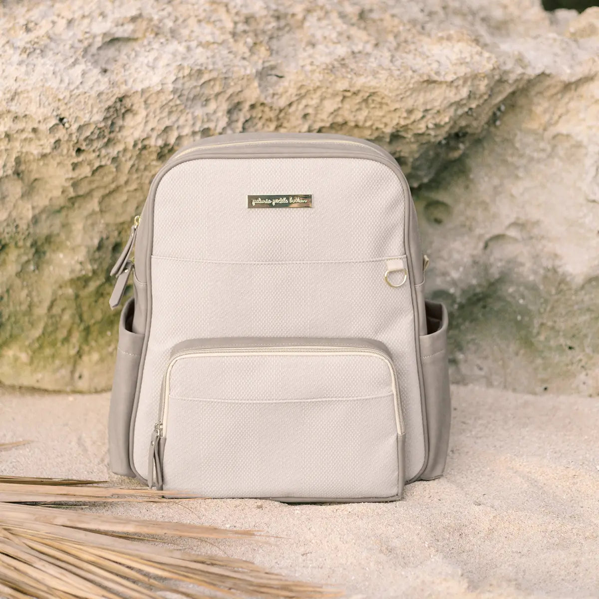 Sync Diaper Bag Backpack shown in grey matte color styled on a sandy beach with rocks in the background