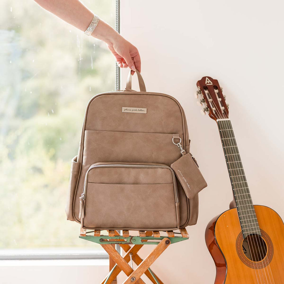 Sync Backpack in Mink shown on bench near window in brightly lit room. accompanied with an acoustic guitar.