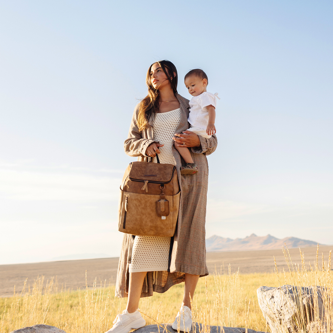 mom holding baby while holding the tempo backpack in brioche with her other hand