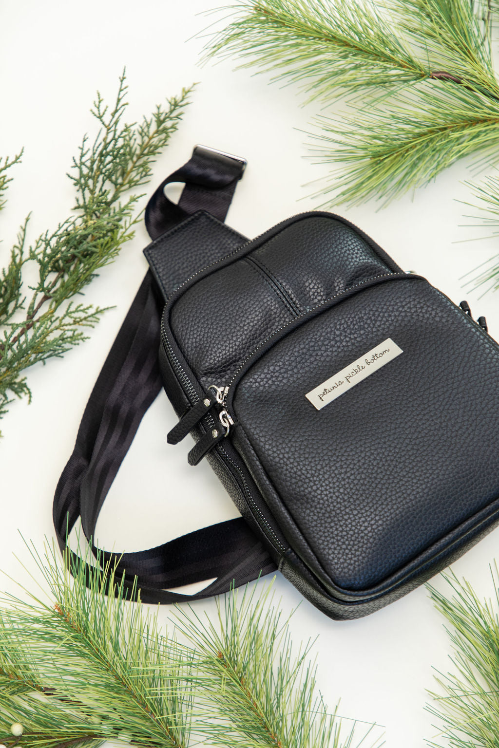 mini criss cross sling backpack in ebony black leatherette with silver hardware styled flat lay with greenery