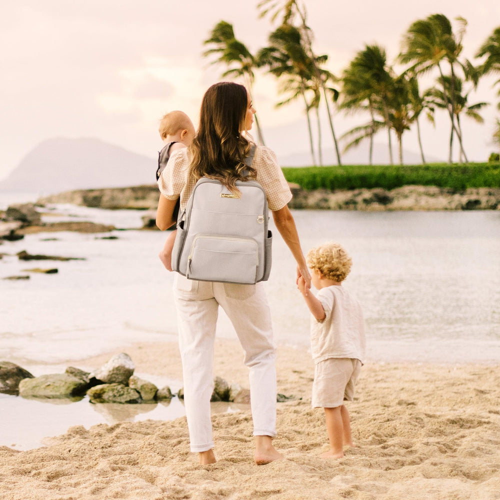 mom wearing sync backpack in grey matte cable stitch while holding baby in arms and holding baby boy's hand